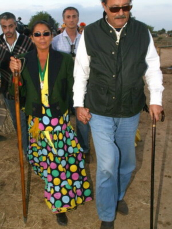 Una pareja camina juntos por un camino rural, sosteniéndose de la mano y usando bastones de caminar.