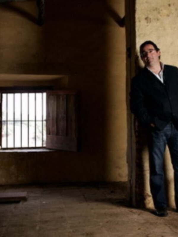 Un hombre se apoya en una pared de piedra, mirando hacia la cámara. La imagen muestra un interior antiguo con una ventana de madera y barrotes, iluminada por la luz natural que entra a través del cristal.