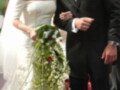 Una pareja casada se besa mientras caminan por un pasillo de boda. La novia lleva un vestido blanco con detalles florales y el novio, un traje negro formal. Ambos sonríen felices en medio de la celebración.