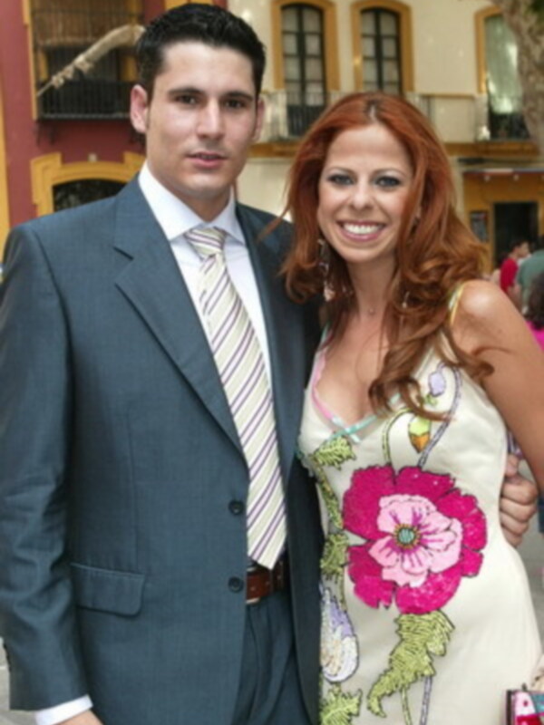 Un hombre en traje y corbata posa junto a una mujer con cabello rojo y vestido floral, frente a una casa con ventanas y una escalera.