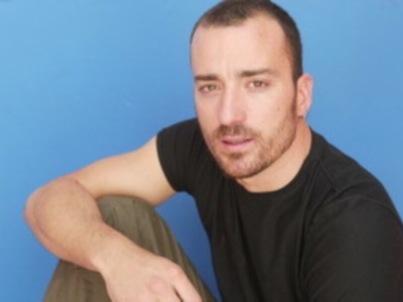 Retrato de hombre joven con camiseta negra, sentado contra un fondo azul.