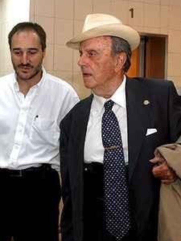 Dos hombres, uno con una camisa blanca y el otro con un traje formal, posando en una sala con una puerta abierta en el fondo.