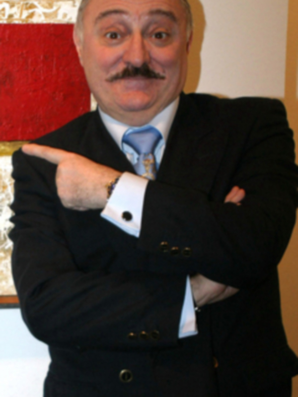 Un hombre con bigote y traje negro, posando con los brazos cruzados en frente de una pintura.