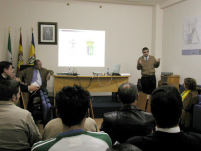 Un hombre en una presentación, frente a un proyector con una imagen de un escudo. La audiencia está sentada en el frente, mirando al orador. El ambiente es formal y parece ser una conferencia o seminario.