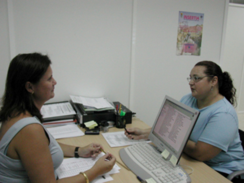 Dos mujeres trabajando en un escritorio con documentos y un ordenador, conversando en un entorno de oficina.