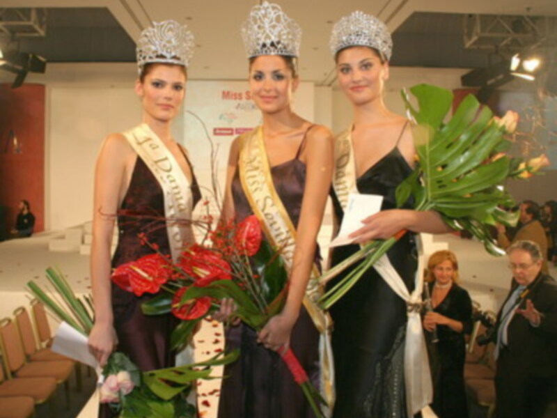 Tres mujeres vestidas con trajes de gala y coronas posan para la foto, una con un ramo de flores.