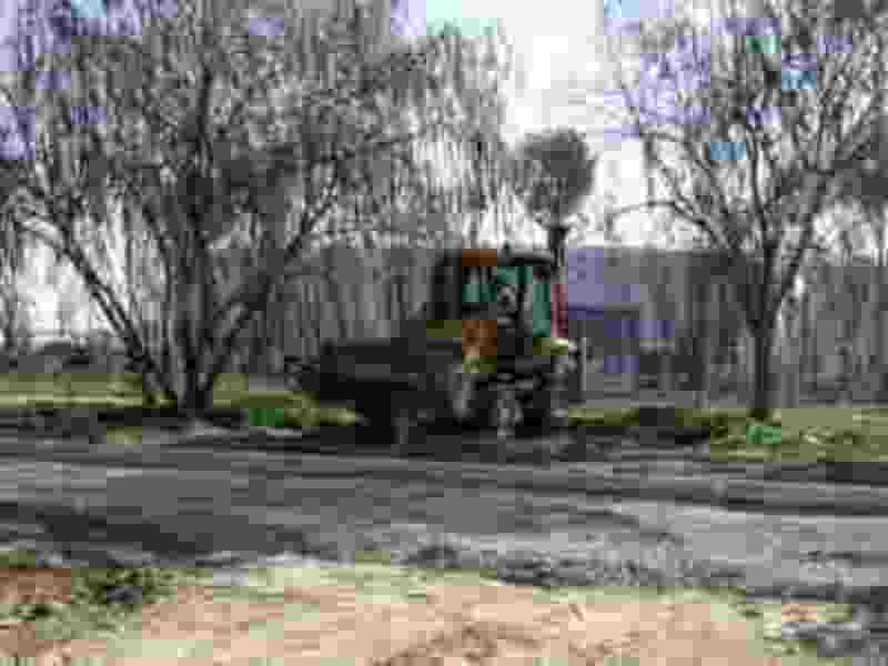 Un tractor amarillo y negro está trabajando en una carretera de asfalto, rodeado por árboles y un edificio en el fondo.