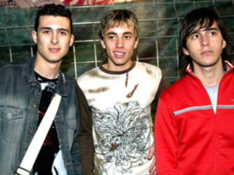 Tres jóvenes posando frente a una pared de azulejos, uno con chaqueta vaquera, otro con camiseta estampada y el tercero con chaqueta roja.