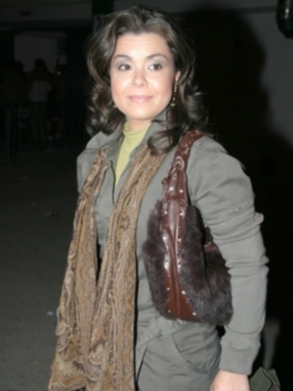 Una mujer con cabello oscuro y rizado, vistiendo una chaqueta de piel marrón y una bufanda de estampado, posa al aire libre en la noche.