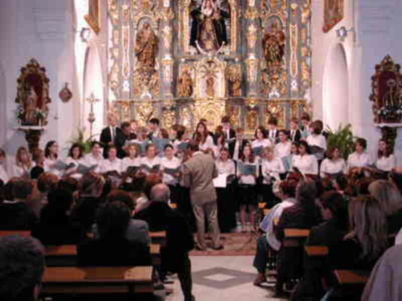 Concierto coral en una iglesia con un coro y un público en el púlpito y bancos de madera.