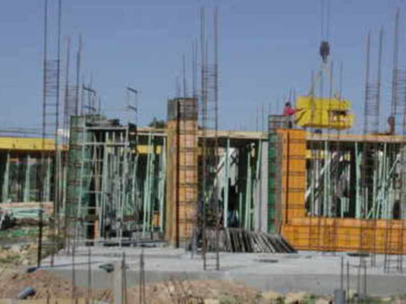 Un edificio en construcción con estructuras de acero y madera, rodeado por pilotes y una cerca. La imagen muestra la fase temprana de la construcción, con muchas columnas verticales y bloques en proceso.