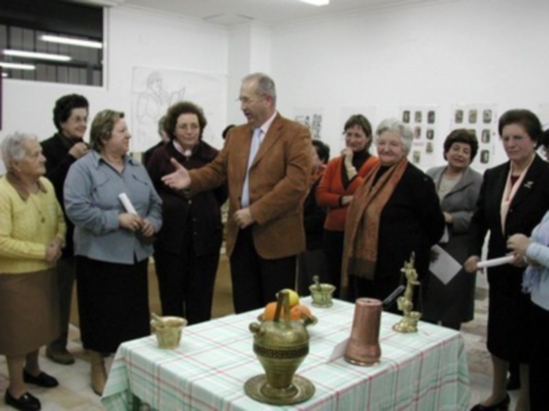 Una sala con personas observando una exposición. En el centro, un hombre hablando a una mujer mientras otras miran atentamente. La mesa tiene un recipiente con flores y documentos. La pared al fondo muestra cuadros.