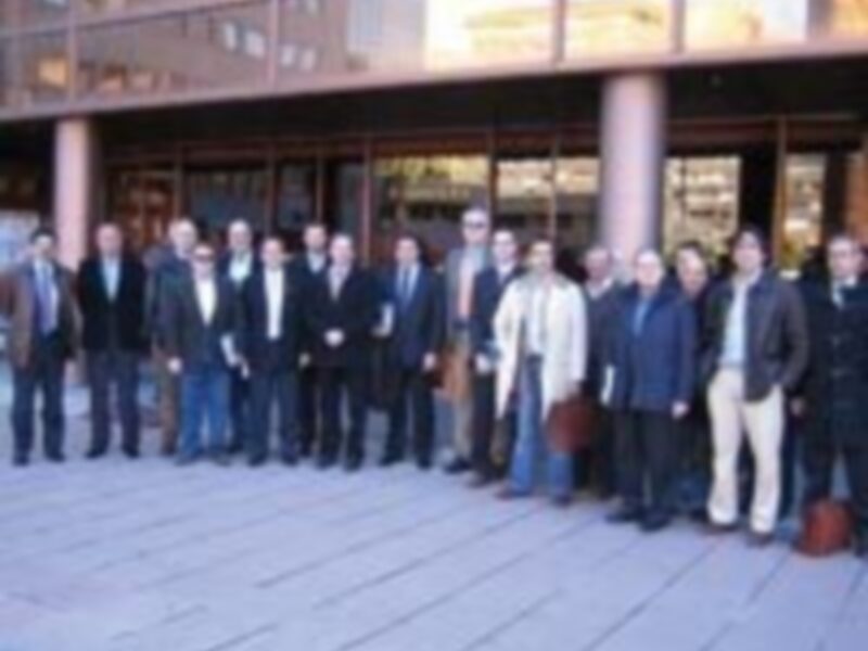 Una foto de una reunión formal con 15 personas vestidas de negocios, posando frente a un edificio moderno.