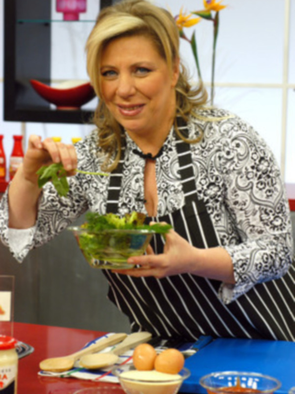 Una mujer con un delantal negro y blanco está sosteniendo una ensalada verde en su mano izquierda. Está sonriendo y parece estar disfrutando de la comida. En el fondo, se pueden ver algunos ingredientes de cocina y un plato con una cuchara.