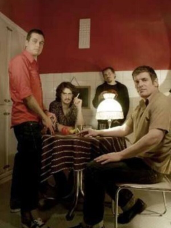 Un grupo de cuatro personas sentadas en una mesa con un fondo rojo y azul. La imagen parece ser de una escena de teatro o una representación.