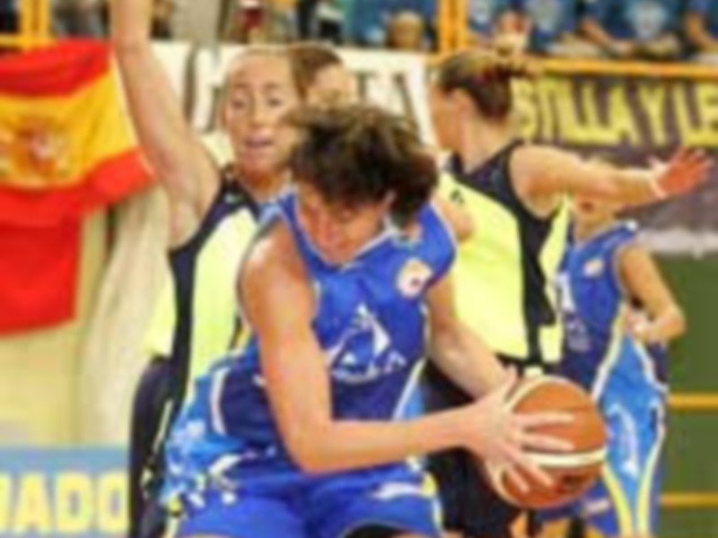 Jugadoras de baloncesto en acción durante un partido. La jugadora del equipo azul sostiene el balón mientras es bloqueada por una jugadora de la otra franja. La bandera española y el escudo del club se pueden ver en el fondo.