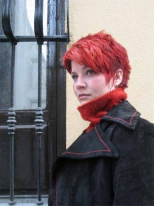 Una joven con cabello rojo corto y una bufanda roja está mirando hacia la cámara mientras se asoma detrás de un portón con rejas.