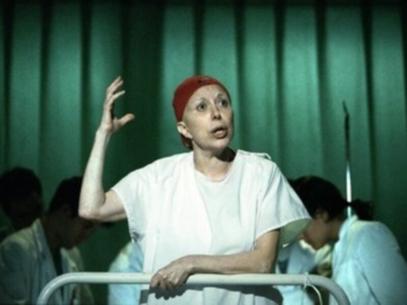 Actriz en escena, vestida con uniforme médico, gesturando con la mano en un entorno teatral iluminado por luces verdes.