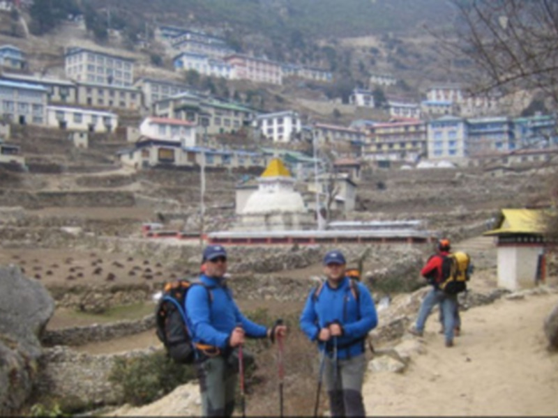 Tres personas con mochilas caminan por un camino de tierra cerca de una ciudad en el Himalaya, con casas y montañas al fondo.