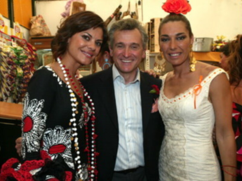 Dos personas sonrientes, una con vestido blanco y la otra con vestido negro y flores rojas, posando en un ambiente festivo.