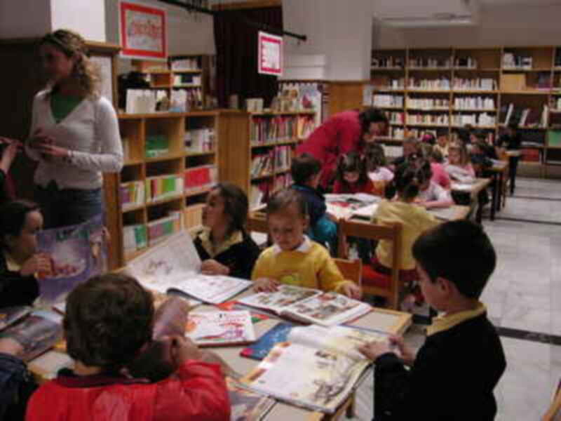 Un grupo de niños en una biblioteca, sentados en mesas y sillas, leyendo libros. La maestra está enseñando a los niños en la parte superior de la imagen. El ambiente es educativo y lleno de libros.