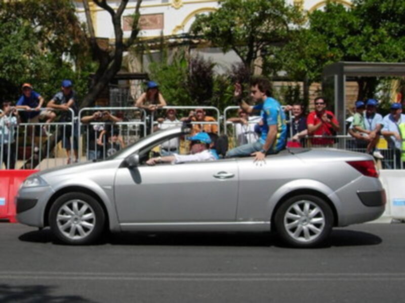 Desfile de coches abiertos con personas saludando a la multitud en una calle con árboles y edificios en el fondo.