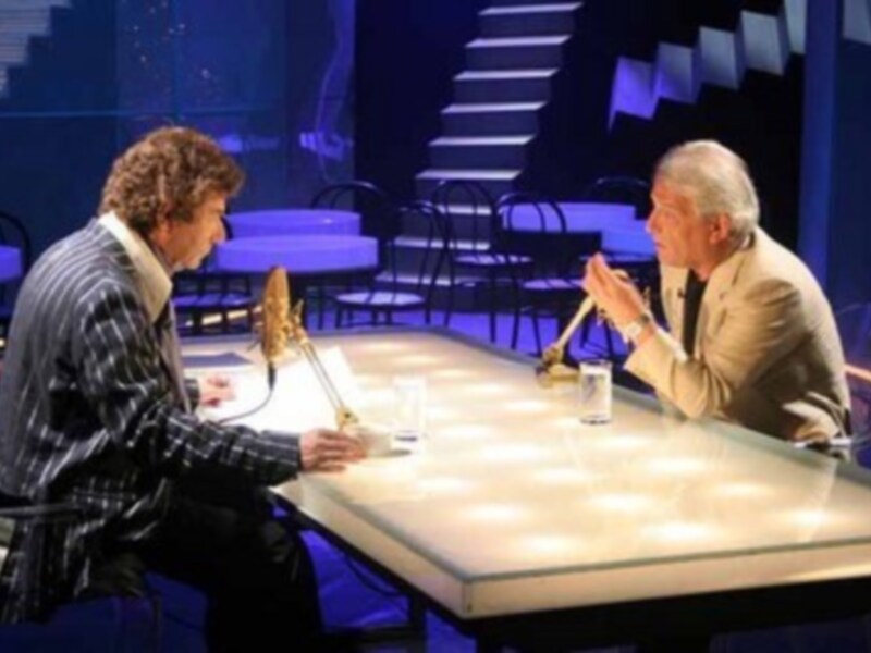 Entrevista en un estudio de televisión con dos personajes sentados en una mesa iluminada.