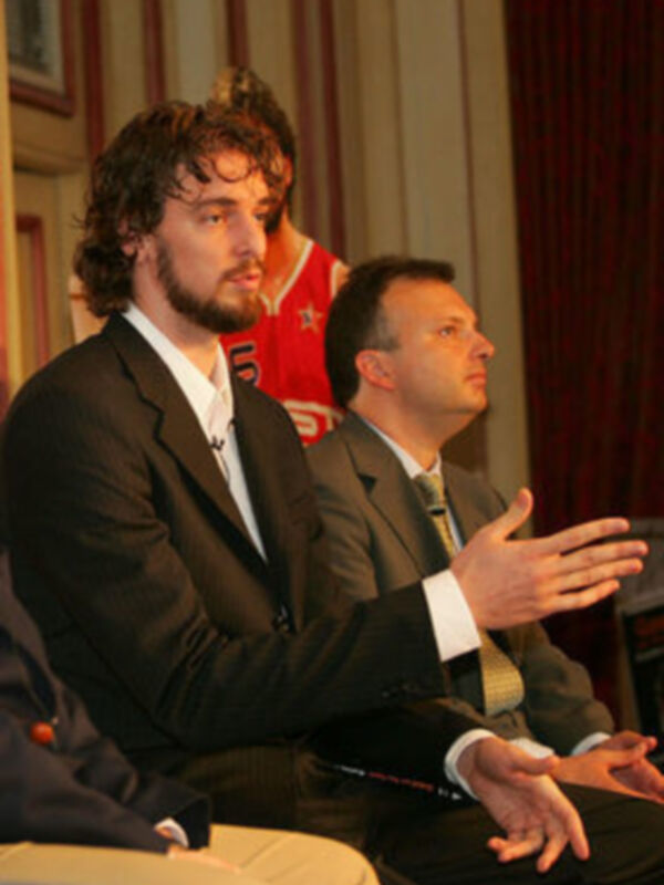 Dos hombres en una conferencia de prensa, uno con una chaqueta negra y el otro con una camisa blanca y corbata, ambos mirando hacia la izquierda.