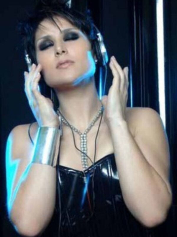 Una mujer con maquillaje intenso y cabello oscuro, posando con los manos en la cabeza. Lleva un vestido negro brillante y una pulsera metálica. La imagen tiene un fondo oscuro con luces de neón azules y blancas.