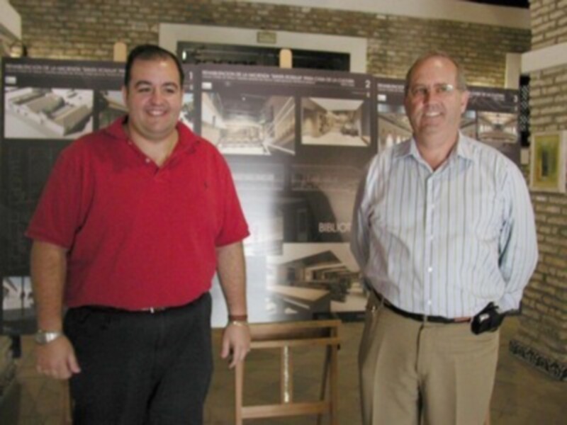 Dos hombres posan frente a un póster con imágenes de interiores, uno en camisa roja y el otro en una camisa a rayas.