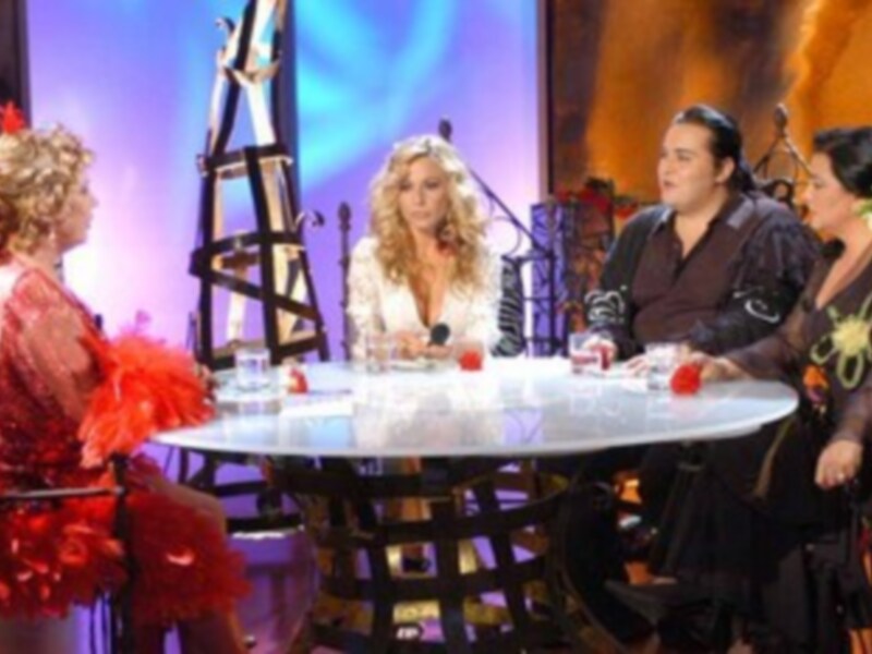 Tres personas en un escenario de televisión, dos sentadas alrededor de una mesa con bebidas y el tercero a su lado. La escena parece ser de un programa de entretenimiento o concursos.