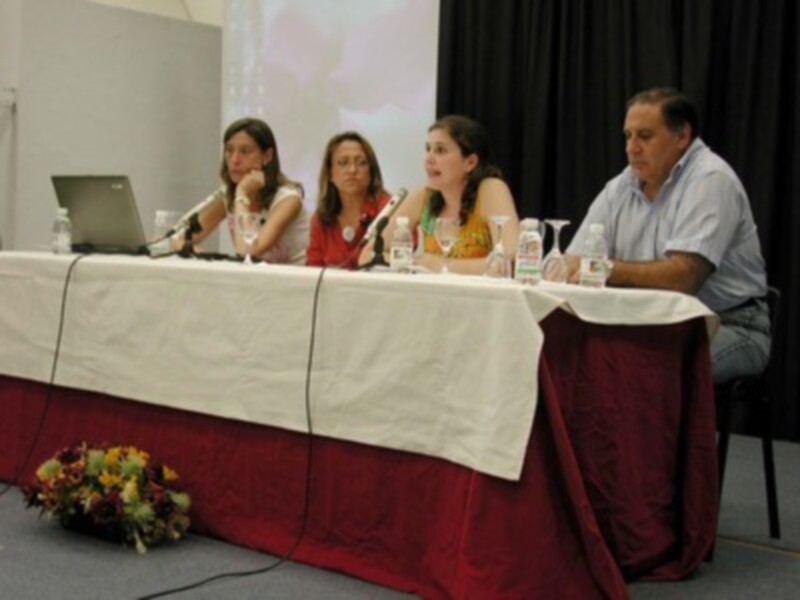 Una mesa con una falda roja y un mantel blanco, cuatro personas sentadas detrás de ella. En la mesa hay dos computadoras portátiles y varios vasos con agua. La imagen muestra a las personas sentadas en sillas, mirando hacia adelante.