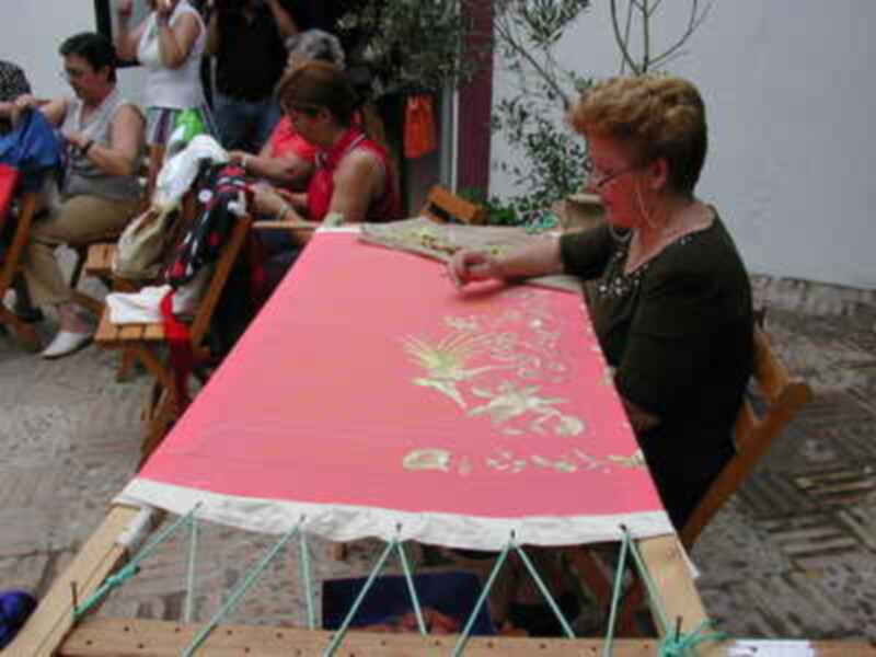 Una mujer está trabajando en un tapiz con motivos florales, mientras otras personas observan. La imagen muestra una actividad artesanal en un ambiente relajado, posiblemente en un taller o mercado.
