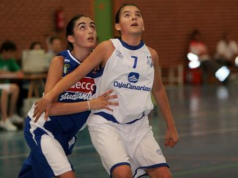 Dos jugadoras de baloncesto en acción durante un partido. Una con el número 7 y otro con una camiseta azul de la marca "CajaCanarias". El fondo muestra un estadio con espectadores y una pantalla de televisión.