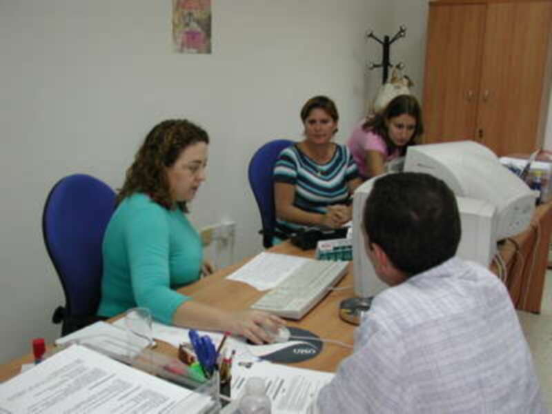 Tres personas trabajando en una oficina, con un hombre frente a un ordenador y dos mujeres sentadas detrás de él. La oficina tiene una estantería, un cartel en la pared y varios escritorios con documentos.