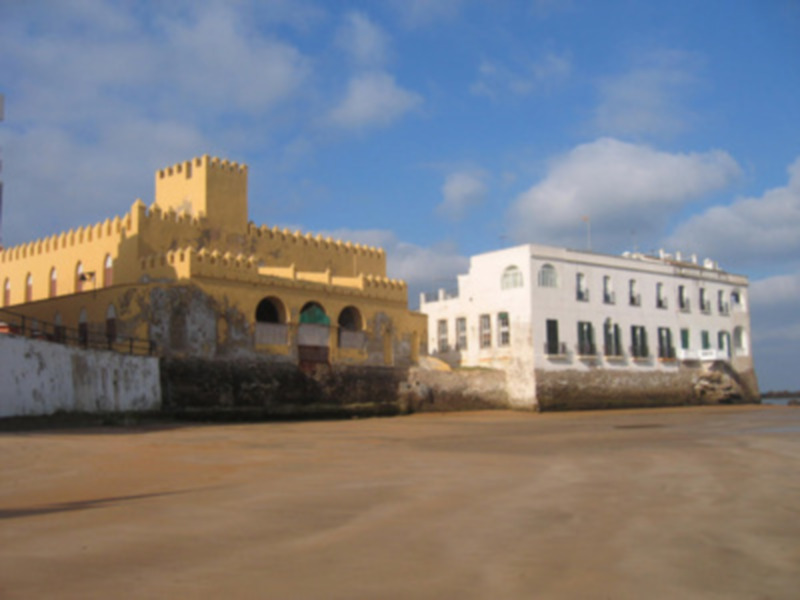 Viejo edificio amarillo con torre en la costa, frente al mar con cielo azul y nubes.