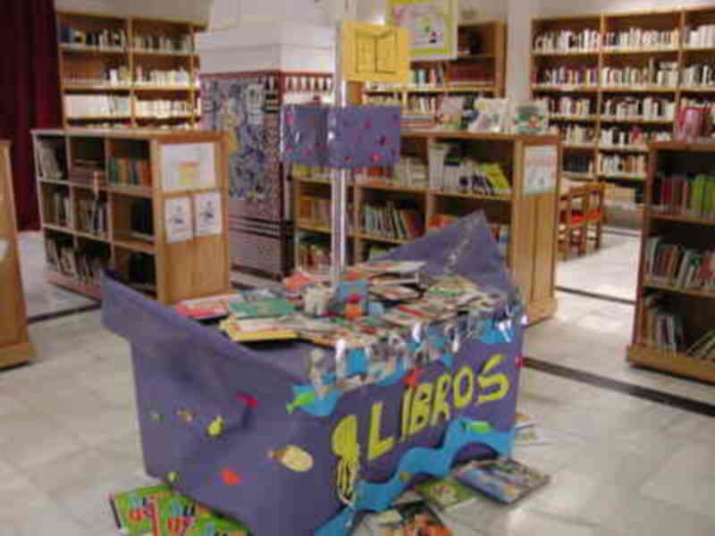 Librería con estanterías llenas de libros, mesita de madera decorada con globos y letras que dicen "Libros", y un cartel con información sobre la biblioteca.