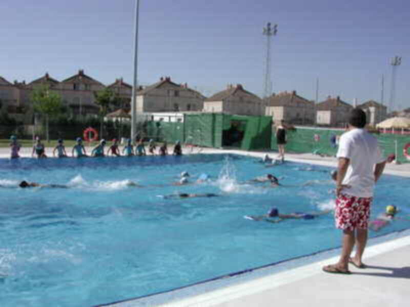 Una piscina al aire libre con niños nadando y adultos observando. En el fondo, edificios de viviendas y un área de juegos infantiles.