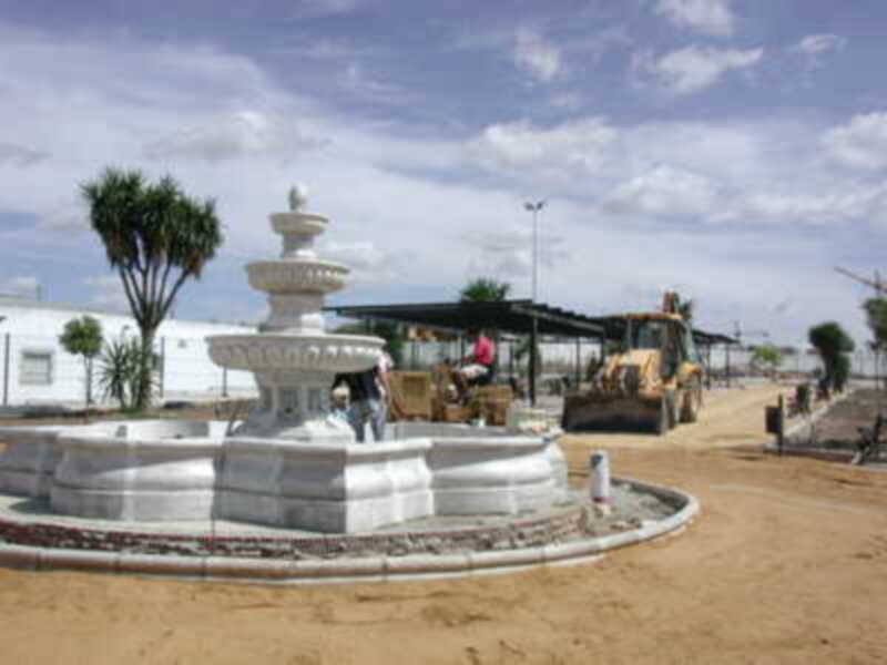 Fotografía de una fuente de agua en construcción, con trabajadores y maquinaria. El cielo es azul con algunas nubes. La imagen muestra un área de construcción en un entorno abierto, probablemente en una ciudad o pueblo.
