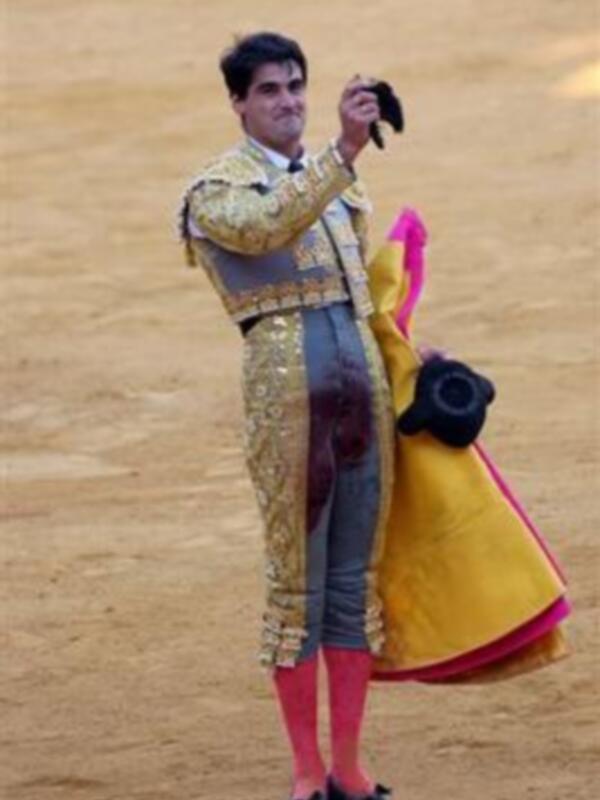 Un torero en traje tradicional de color dorado y rosa, con una espada en la mano izquierda, posa sobre un fondo de arena.
