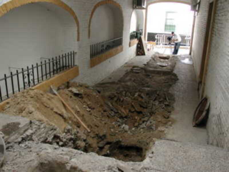 Un antiguo edificio con paredes blancas y arcos amarillos está siendo restaurado. Se pueden ver restos de piedra y tierra en el suelo, mientras que una persona está trabajando con herramientas cerca de un toldo.