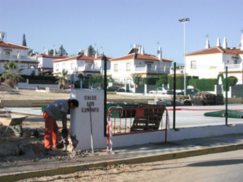 Un trabajador está colocando un letrero en una pared blanca con letras negras. El letrero dice "Calle Los Limones". En el fondo, se pueden ver casas blancas con tejados rojos y un área de construcción.