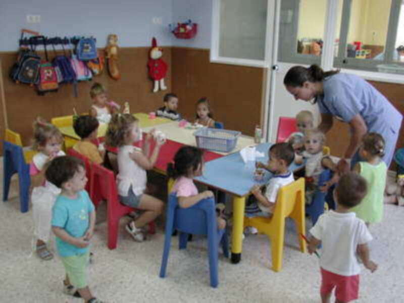 Niños pequeños jugando en una guardería con sillas de colores y mesas. Una niña está tomando un vaso cerca de una mesa con juguetes y libros. La guardiana está observando a los niños.