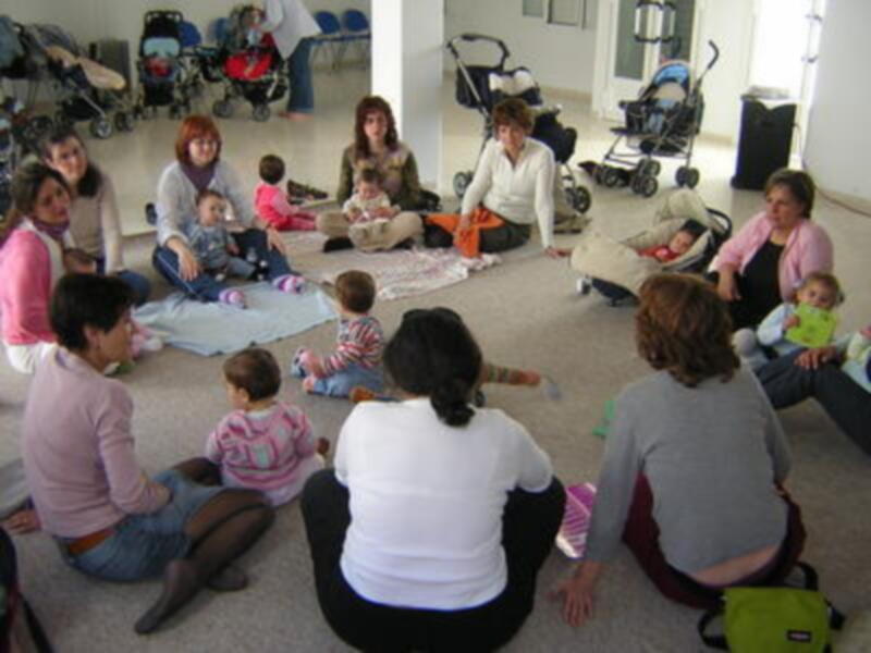 Niños y adultos sentados en el suelo, con carros de bebé alrededor.