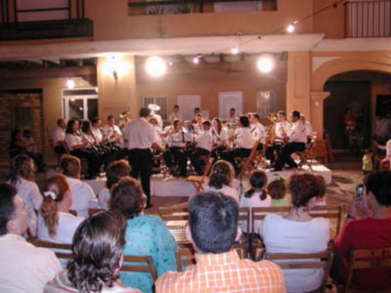 Una banda musical toca en un escenario mientras una audiencia de personas observa la actuación. La sala tiene una decoración simple y un ambiente festivo.