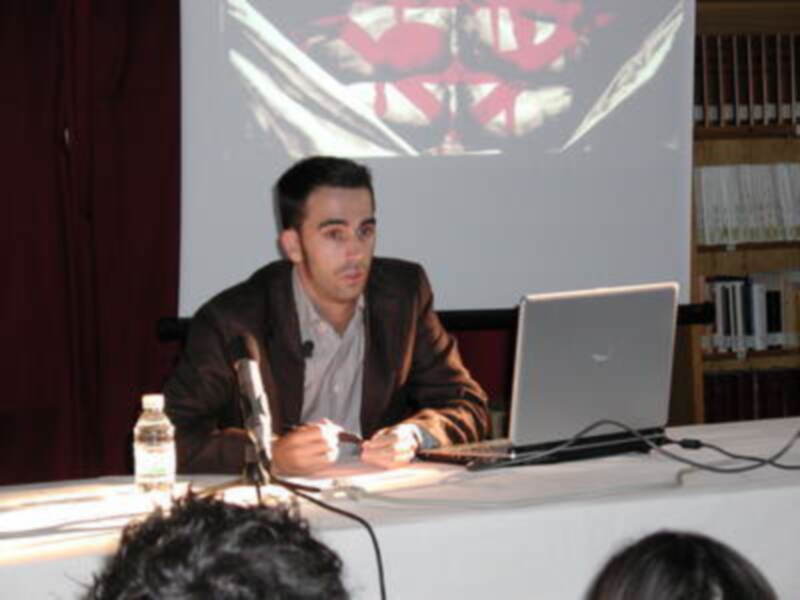 Persona hablando en un evento, con un proyector en la pantalla y un ordenador portátil en la mesa.