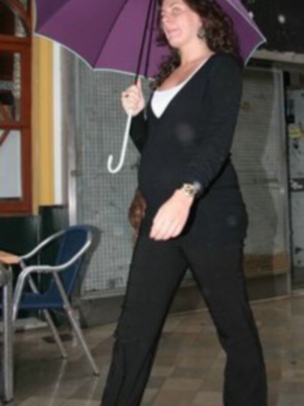 Una mujer camina con un paraguas púrpura en un ambiente interior, vestida con un traje negro y blusa blanca.