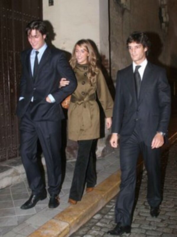 Tres personas caminando juntas en una calle adoquinada, vestidas formalmente con trajes y abrigos.