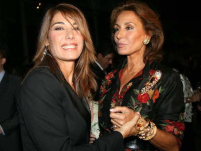 Dos mujeres sonrientes en un evento formal, una con vestido negro y la otra con un vestido floral negro.
