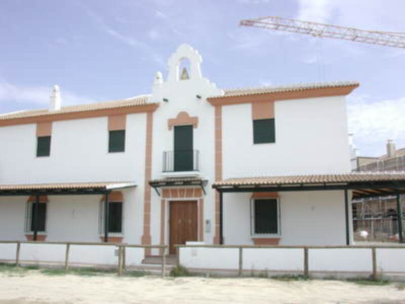 Casita blanca con detalles en terracota y balcones verdes, ubicada en una zona de playa con arena y madera.
